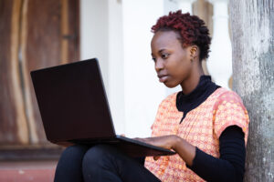 Concentrated African teenager sitting on the porch with her laptop on her legs preparing online for her high school exam; education during COVID pandemic lockdown, e-learning